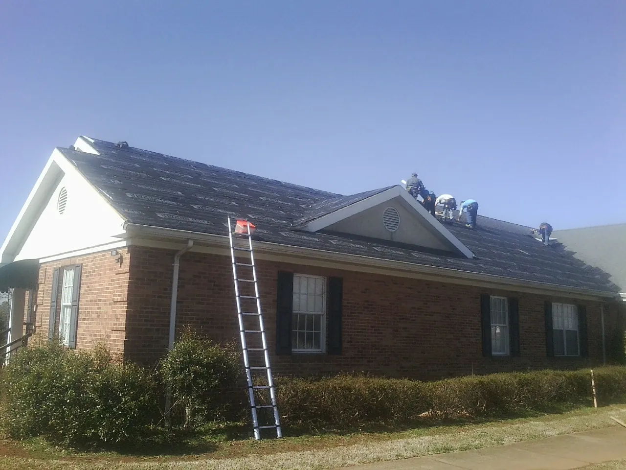 Crew tearing shingles off a brick ranch roof with a ladder set — Emergency Roof Repair work by our crew in Bonneau