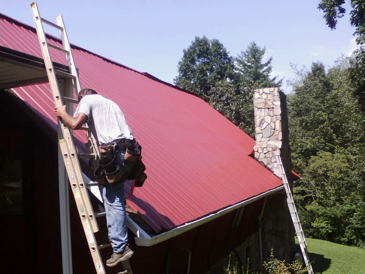 Roofer on a ladder beside a red metal roof and stone chimney — Soffit & Fascia Repair by Roofing Brothers in Bonneau